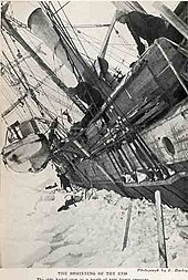 Side of a wooden steamship held in solid ice, leaning steeply to the left with a lifeboat swinging in its davits. One man visible on the ice, another aboard the ship, looking down.