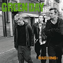 A black-and-white photo of the band members walking along a sidewalk. On the top left, the band name is written in green, and on the bottom right, the album title is written in yellow as "WARNING:".