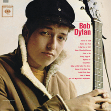 A close-up of Bob Dylan wearing a coat and hat, holding a guitar