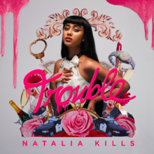 A young, tan-skinned woman wearing a white dress with her hand on her chest standing against a grey background. Around her are various items including watches, champagne, police cars and roses. The word "Trouble" appears on the centre of the image. The text "Natalia Kills" appears at the foot of the image. Pink liquid flows down from the upper corners of the image.