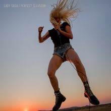 Gaga singing into a microphone while jumping. She wears shorts and a T-shirt with her hair in a ponytail. Behind her, a sunset can be seen.