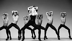 Black-and-white image of Madonna wearing tight black dress dancing in high heels. Behind her, a group of bare-bodied men emulate the moves, wearing similar high-heeled shoes