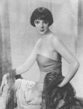 young, slim white woman with straight dark hair in low cut evening gown, seated and turning her head to look at the camera
