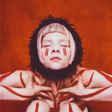 Aurora is against a reddish background with her eyes closed and hands touching her chest. The subject has red marks in her skin and a black scarf around her neck.