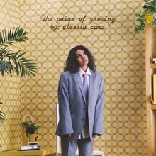 Alessia Cara, wearing a grey suit, stands afront a cream-coloured background with the album title and her name displayed above her