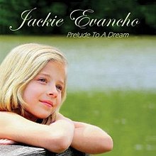 A photo of nine-year-old Jackie Evancho gazing over a grassy area and resting her crossed arms on a wooden fence railing. A lake and wooded area are in the background.