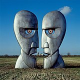 A colour photograph of two large silver-grey iron sculptures of opposing silhouetted faces. The sculptures are standing in a brown wheat field with a blue sky behind them.