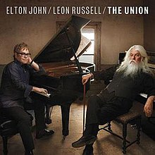 In a room, Elton John and Leon Russell (holding a staff) sit on piano benches while next to pianos. A window is seen behind a piano with lighting shining out into the room.