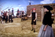 A man wearing a black turtleneck and a red hat holds a whip while a woman in a black dress smoking a cigarette with a holder stands in front of him. In the background are onlooking cowboys and cowgirls, four men wearing black turtlenecks playing instruments, and a ranch house.