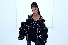 Ariana Grande standing, her hair in a ponytail, wearing black-oversized jacket and boots as she is staring at the camera.