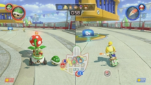 Two characters in split-screen view on a concrete surface. On the left is Bowser Jr., a yellow turtle-like creature with a spiked green shell who is riding in a small circular car. In front of him is a Piranha Plant, a venus flytrap-like creature with a white-spotted red head. On the right is Isabelle, a humanoid yellow dog on a motorbike. Isabelle is driving towards a blue switch with a key icon placed below a yellow and blue cage. In the center of the screen is a minimap displaying the characters' location in the course.
