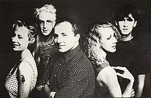 Five adults framed in upper body. In the front row, the left female is shown in left profile, slightly turned to her left and smiling, she wears a white dress with black polka dots and has a serpent tattoo on her upper left arm. Behind her, a slightly balding male is more turned towards the front and has his arms folded across his chest, his shirt is dark with white polka dots. Back to back to him is the second female in right profile with her right hand touching her shoulder. In the back row, the left male has white hair and is facing forward, he is wearing glasses and has an obscured design on his shirt. The right male has dark hair, he is staring forward and wears a black tee shirt.