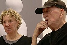 An elderly man and woman dressed in black, with art gallery background. She has dyed, blonde curly hair; he has a baseball cap.