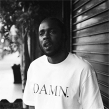 A black and white image of Kendrick Lamar standing next to a closed storefront dressed in a white shirt with the text "DAMN." printed on the shirt.
