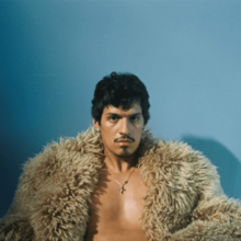 Omar sitting partially bare-chested, wearing a furry jacket and a cross necklace, looking ahead at the camera in front of a blue background.