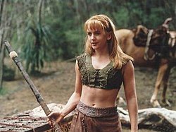 A young blonde woman wearing a green woven tank-top and leather skirt. She holds a wooden fighting staff; a horse can be seen in the background.