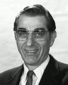 Clean-shaven, bespectacled man, in his 50s or older, with oiled black hair, wearing suit and tie.