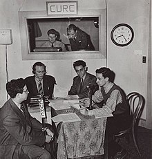 Four people are seated around a table in a radio studio. Two others watch them rom a window. There is a banner with the letters "CURC" above the window.