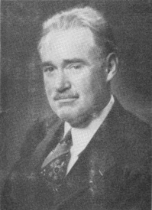 Monochrome portrait, from the chest upwards, of Bishop, probably in his fifties. His hair is neatly combed back from a somewhat receding hairline. He has a neatly trimmed moustache. He wears a dark jacket, a light-coloured shirt, and a patterned tie.