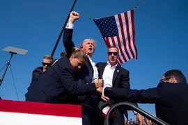 A man with his fist held above his head, blood is on his ear and cheek, the American flag flutters in the air behind him, and two federal agents shield and escort him