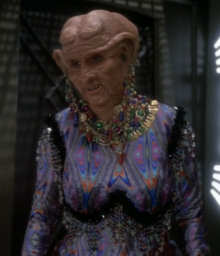 A Caucasoid woman in the heavy orange makeup and prostheses typical of Ferengi looks to the camera's left; she is wearing a garish, skintight outfit and excessive jewelry from her clothes, neck, and lobes