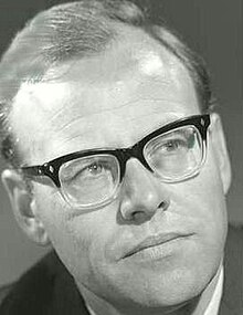 Publicity headshot of clean-shaven man with pale, oiled hair, brushed back, and spectacles.