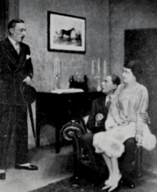 Smartly dressed young white man entering a room where a young man and women appear to be intimately seated