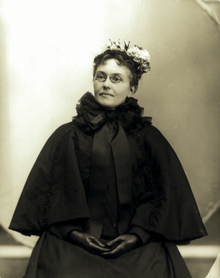B&W portrait photo of a seated middle-aged woman wearing a dark cloak and gloves, as well as flowers in her hair.
