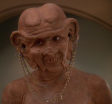 A Caucasoid woman in the heavy orange makeup and prostheses typical of Ferengi looks to the camera's left; she is nude, baring her upper chest and shoulders