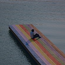 Baby Keem sitting on a rainbow colored wooden pier surrounded by water.