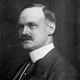 A photographic portrait of a man, showing his head and shoulders. He has a moustache and dark, combed back hair. He wears a jacket, a shirt with a high imperial collar and tie.