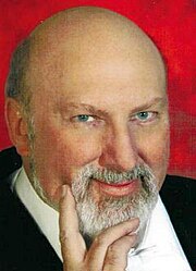 Three-quarter view, publicity shot of a bald, smiling man with neat, white beard and blue eyes.