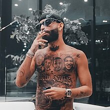 A shirtless man with tattoos and wristwatches is seen smoking.