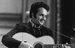 A dark-haired man wearing a dark jacket and playing a guitar