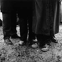 Black and white photo with people's trousers and shoes standing on dirt.