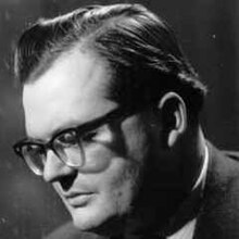 Profile publicity shot of a clean-shaven man with short, dark hair and spectacles, in dramatic pose, looking down.