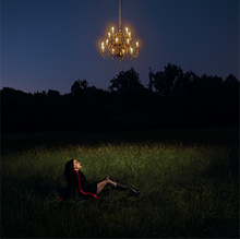 The cover depicts a woman sitting in a darkened field; above her head, hanging from the sky, is a chandelier