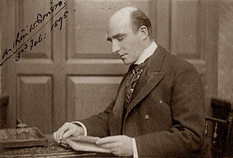photograph of middle-aged white man, bald, clean-shaven, sitting at writing-desk