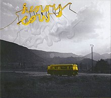 A black and white photograph of a road with mountains in the background; on the road is Henry Cow's touring bus coloured yellow; at the top of the image over the sky are the letters "Henry Cow" in yellow reflecting on the clouds