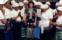 Image of a group of several women feeling reflective. A white woman with dark hair is in the middle, wearing a black dress, holding hands with some of the women, who are black and wearing white outfits and mobcaps.