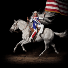 A woman with long white hair sits reverse side-saddle atop a white horse. Her cowboy outfit is in the colors of the American flag, which she holds, only the red and white stripes visible.