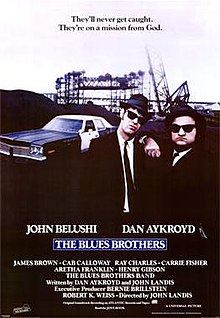 Movie poster with two of the main characters on the right side of the image: They are both wearing black suits, hats, and sunglasses and facing forward. The man on the right is resting his arm on the shoulder of the man on the left. A police car is present on the left side of the image behind them. At the top of the image is the tagline, "They'll never get caught. They're on a mission from God." At the bottom of the poster is the title of the film, cast names, and production credits.