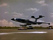 A blue, futuristic airliner rests on top of three orange support vehicles (one underneath the fuselage and two underneath the wings) as it speeds out of control down an airport runway, the sky bright with clouds.