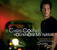A man stands in front of an out-of-focus sports car. The background is a gradient that goes from red to green. Besides the man is written "Chris Cornell", with "You Know My Name" below it. In the down right corner is the text "As featured in the motion picture CasinO ROyale", with a gun-like "7" below Royale's "O".