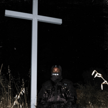 A night scene outside in which a large white cross looms above JPEGMafia, who is covered head-to-toe in black.