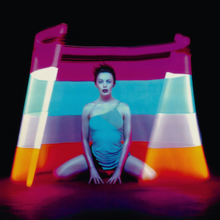 A body image of a young woman (Kylie Minogue) inside a cut cone with multi-coloured lights. From top: Purple, blue, mid-purple/blue, oranges and red. The woman is wearing a small blue mini dress with detail on the top left, whilst the floor has a reflection of the cone.