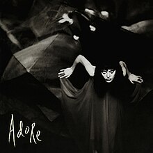 A black-and-white photo of a Caucasian woman leaning forward while holding the ends of a flowing black dress. In the corner, "Adore" is displayed in white handwriting.