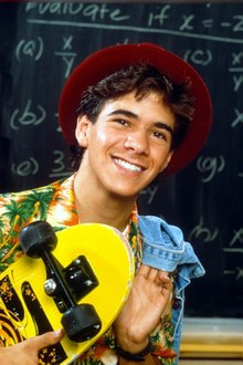 A teenage boy wearing a red fedora and a Hawaiian shirt smiles to the camera while holding a yellow skateboard in his left hand and a denim jacket over his right sholder