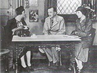 Middle-aged white woman in smart 1930s costume addresses weary-looking man and dazed woman across a café table