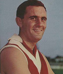 A dark haired white man smiles off camera while wearing a red woolen football jumper featuring a white chevron